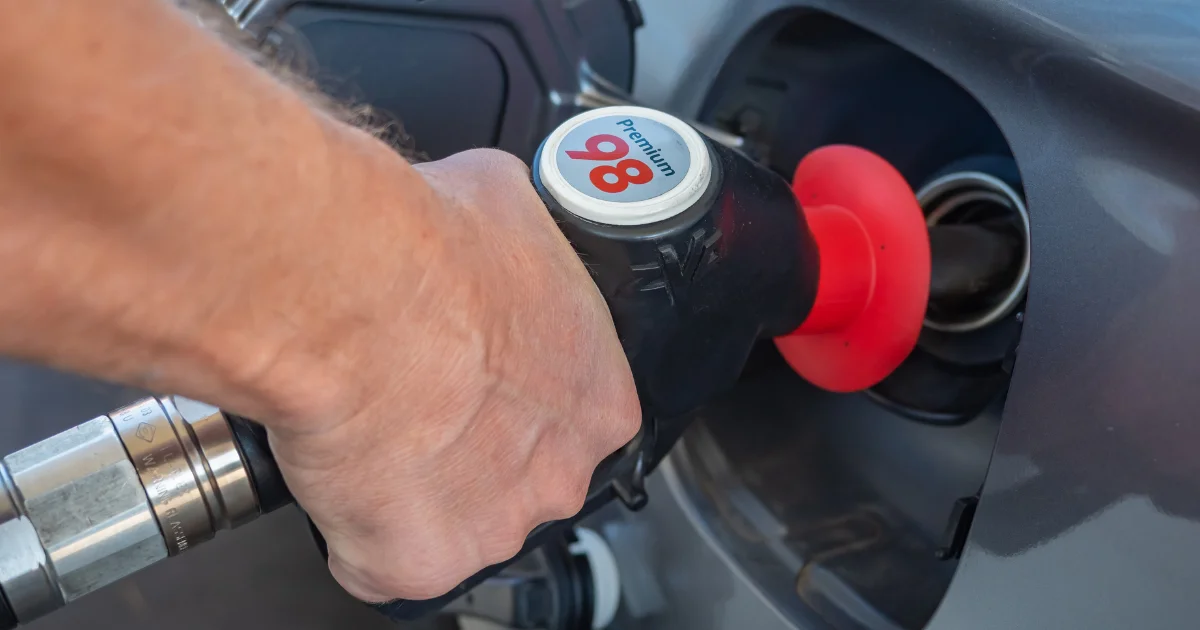 A close-up of a person's hand holding a black and red fuel pump nozzle labeled "Premium 98" while inserting it into a car's gas tank to fill up on petrol.