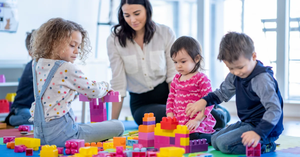 A female childcare educator watching over three young children as they sit on a colorful play mat and build towers out of large, colorful plastic toy blocks.