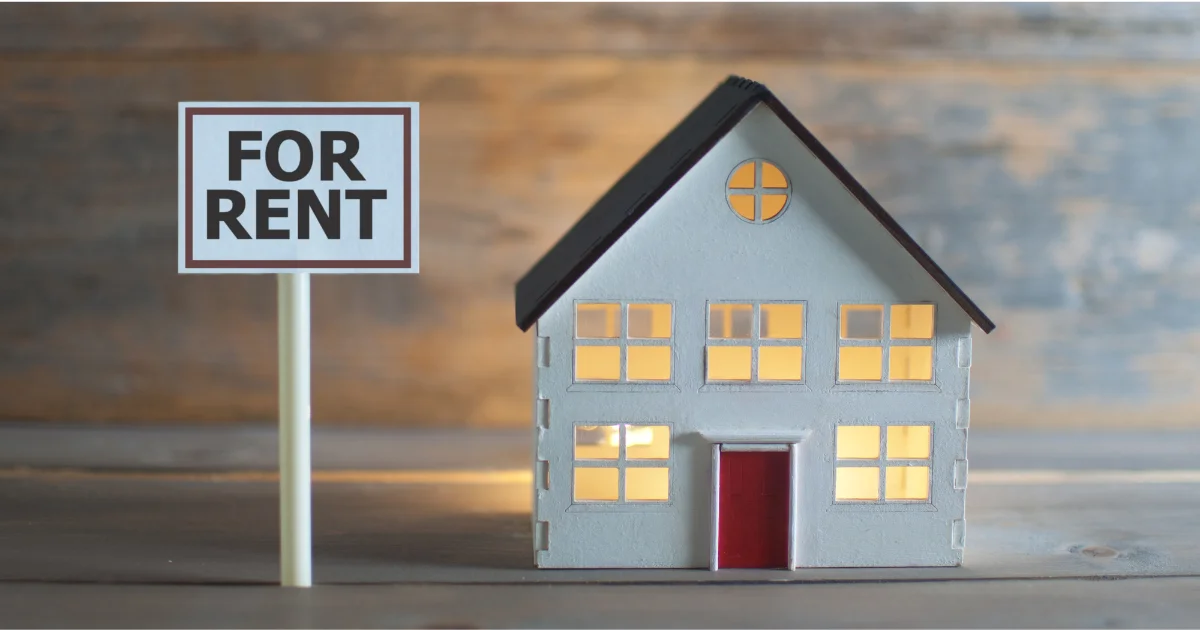 A small white model house with glowing windows sitting next to a miniature sign that reads "FOR RENT" against a rustic wooden background.