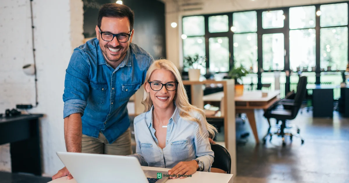 A smiling man and woman, both wearing glasses, pose by a laptop in a modern co-working office space.