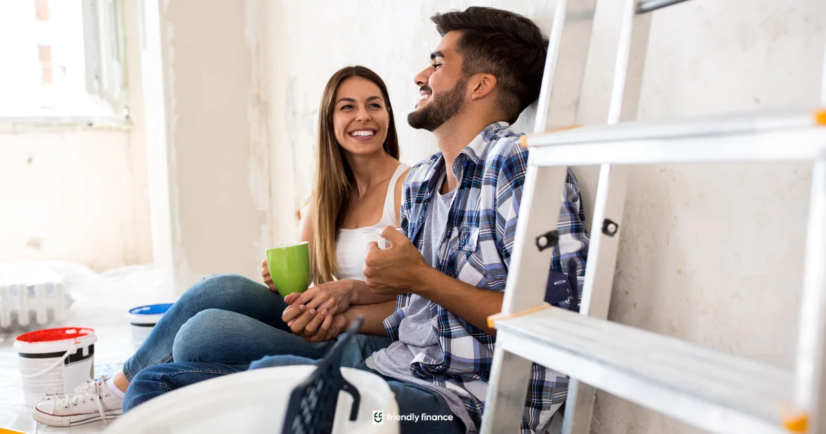 A cheerful young couple sits on the floor of a room under renovation, holding coffee mugs and laughing, with a ladder and paint cans nearby.