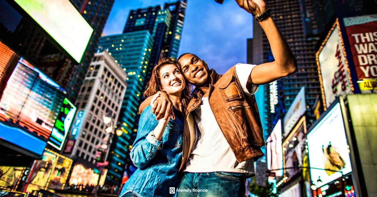 A happy couple takes a selfie together at night in Times Square, New York, with bright city lights and billboards in the background.