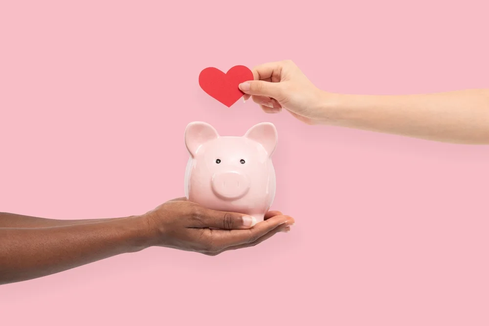 Two hands holding a pink piggy bank with a heart being placed inside against a pink background