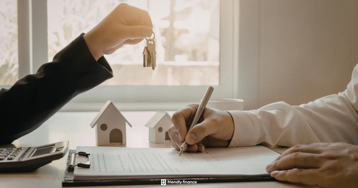 A close-up of a person signing a contract while receiving a set of house keys. Small white model houses sit on the desk, representing a mortgage or home loan.