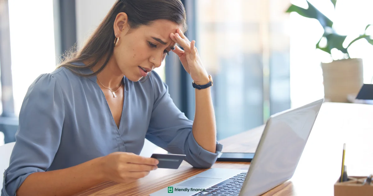 A worried woman holds a credit card to her head while looking at her laptop screen, appearing stressed about her finances or online banking.
