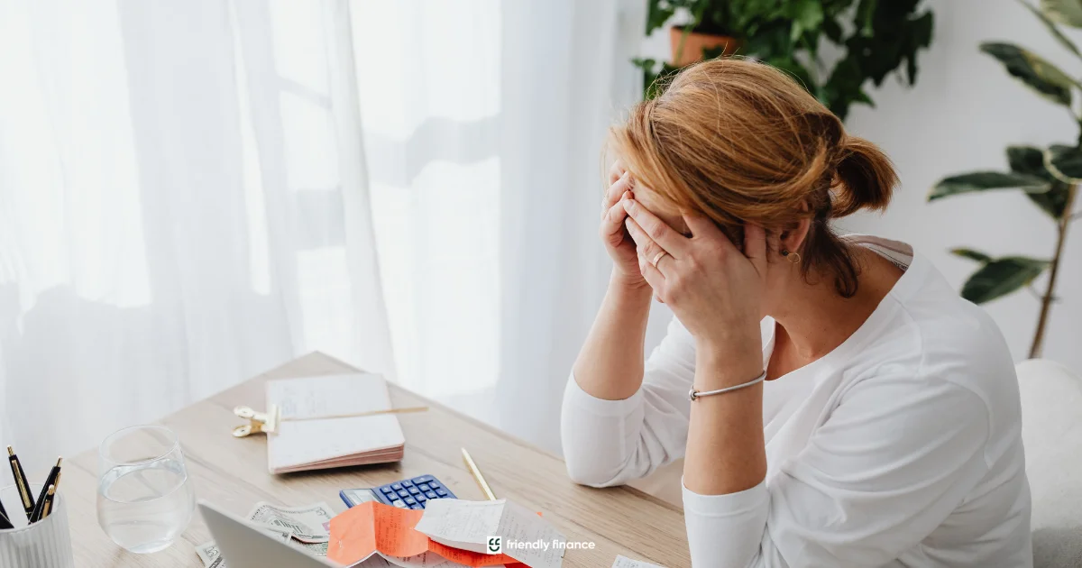 A stressed woman sits at a desk with her head in her hands, surrounded by a calculator and unpaid bills, illustrating financial worry or debt.